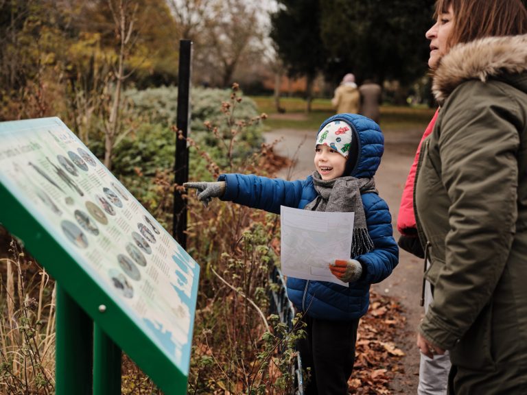 Small child dressed in outdoor clothing reads an information board about nature, smiling and pointing.